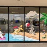 Colorful beach-themed dog mural painted for Canine Kingdom (formerly Beach For Dogs), featuring four playful cartoon-style dogs: one digging, two playing with a beach ball and a frisbee, and one floating on water. Includes whimsical elements like a fire hydrant sandcastle and a salmon-shaped palm tree. Painted in a bright, animated, and fun style suitable for a dog daycare or pet business