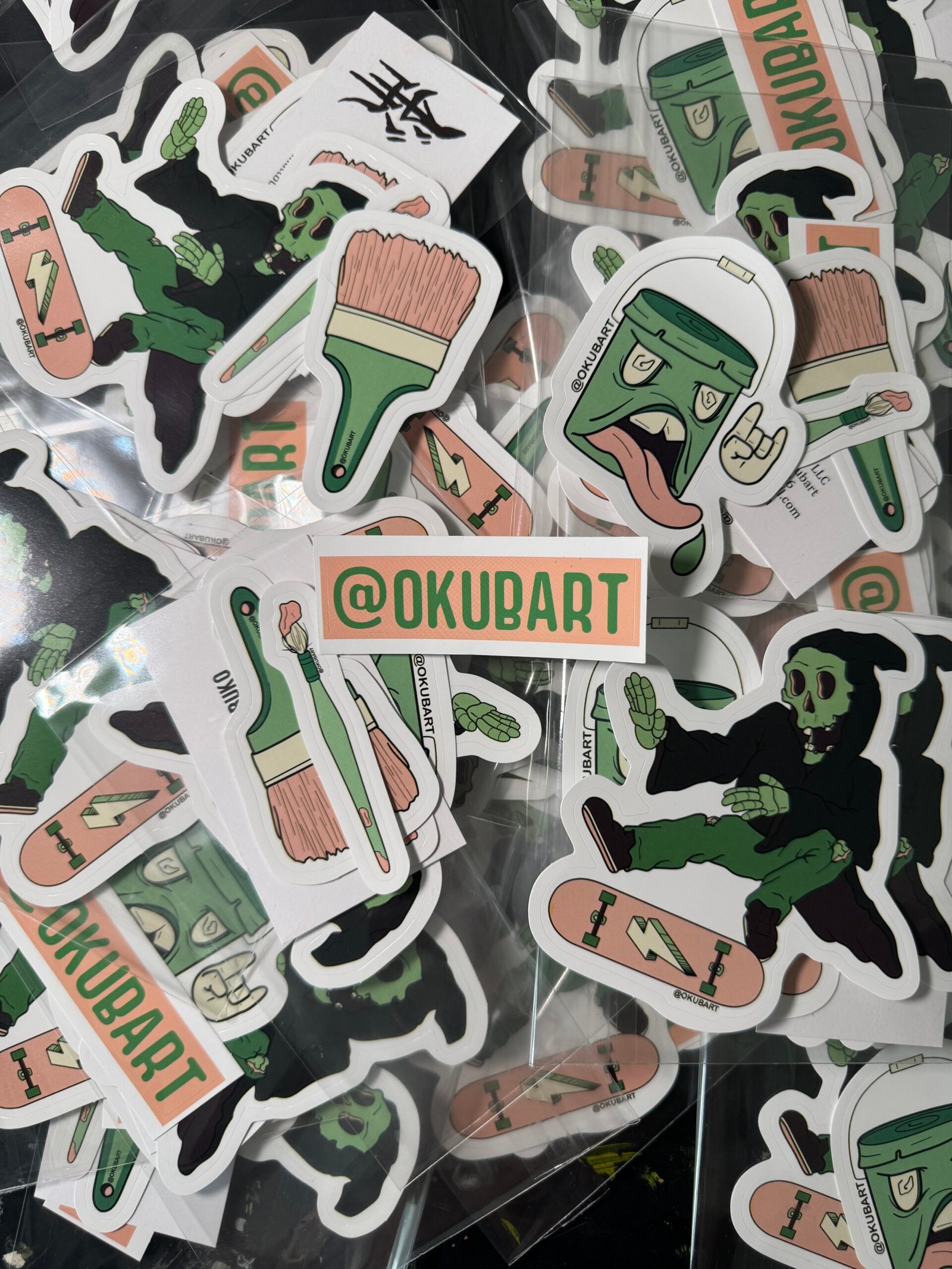 Five-piece vinyl sticker pack featuring bold, cartoon-style artwork: a five-gallon paint bucket with a face, two expressive paintbrushes (thin and thick), a dynamic skeleton or grim reaper doing a kickflip on a skateboard, and an @okubart artist tag. Designed in a colorful, street art-inspired style with bold outlines and graphic flair.